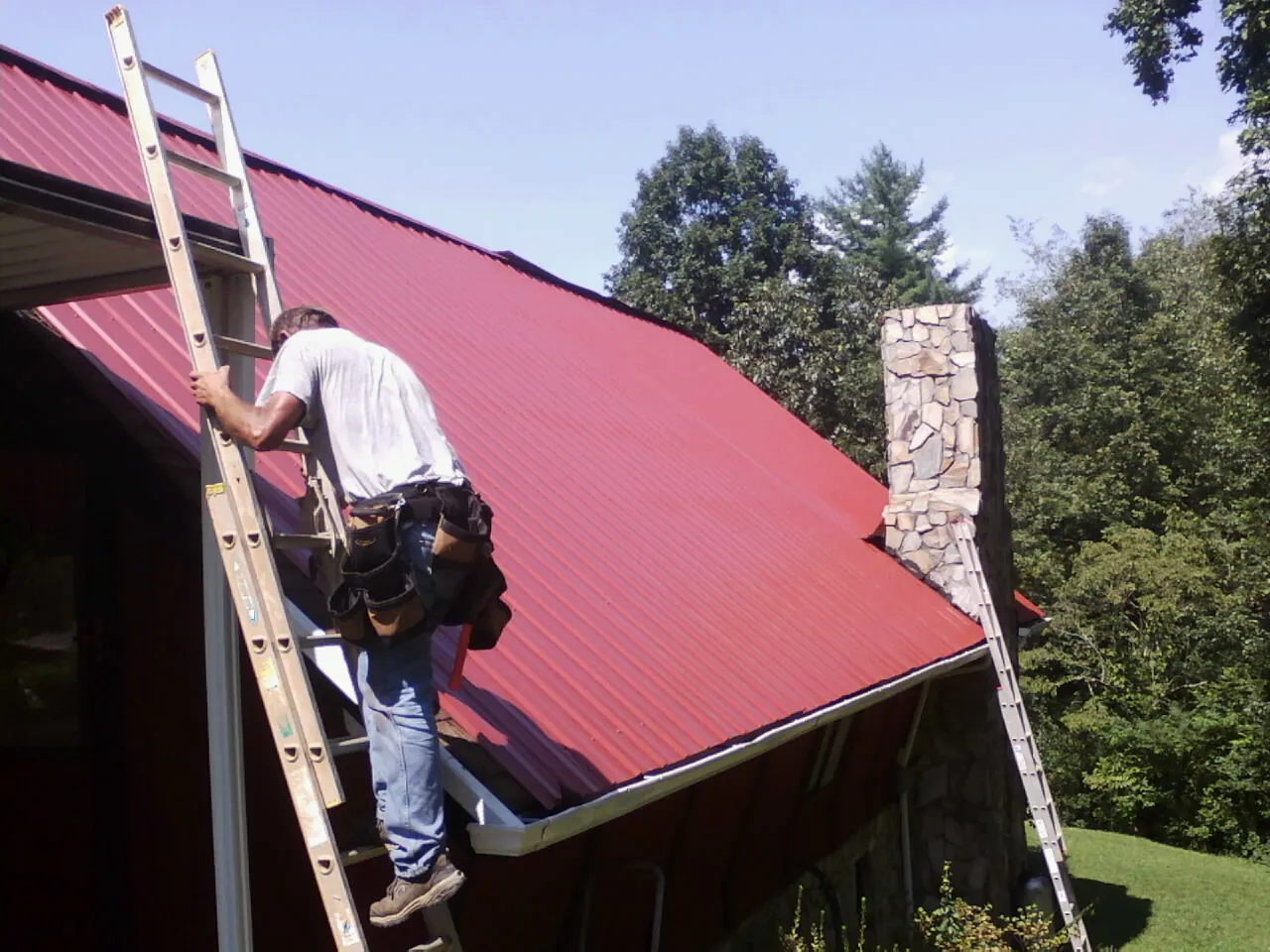 Roofer on a ladder beside a red metal roof and stone chimney — Soffit & Fascia Repair by Roofing Brothers in Costa Mesa